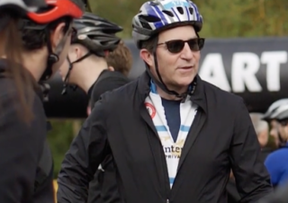 a man wearing an Advent International cycling jersey , sunglasses, and a bike helmet standing in a crowd of other cyclists